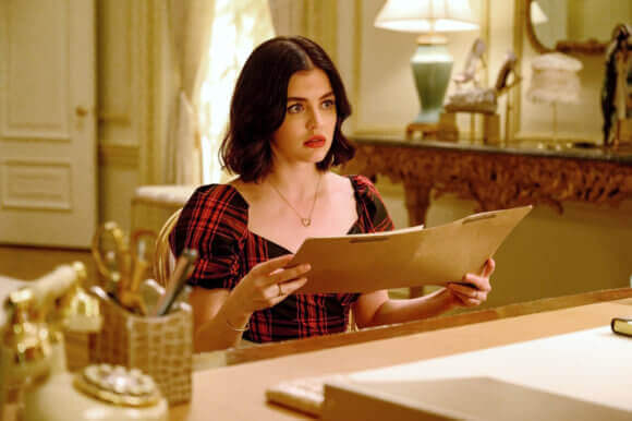 katy Keene - a brunette woman with dark red lips and wearing a red plaid dress with poofy sleeves - holds a document in her hand in a sunlit office. She is looking up at the person sitting across from her and unseen by the camera with incredulity.