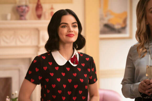 Katy keene, a brunette woman, wears a black, short-sleeved dress dotted with red hearts and a white peter pan collar. She smiles toward someone off-camera in a sunny, well-lit room at Lacy's department store