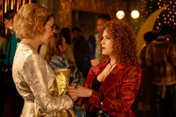 Pepper Smith, an asian-american woman with short, curled blonde hair and glasses, who is wearing a silver jacket, holds hands with Miss Freesia, an older woman with wild red shoulder-length hair, a crushed red velvet jacket, and a black, low cut top. Miss Freesia holds Pepper's hand and looks at her with concern in her eyes.