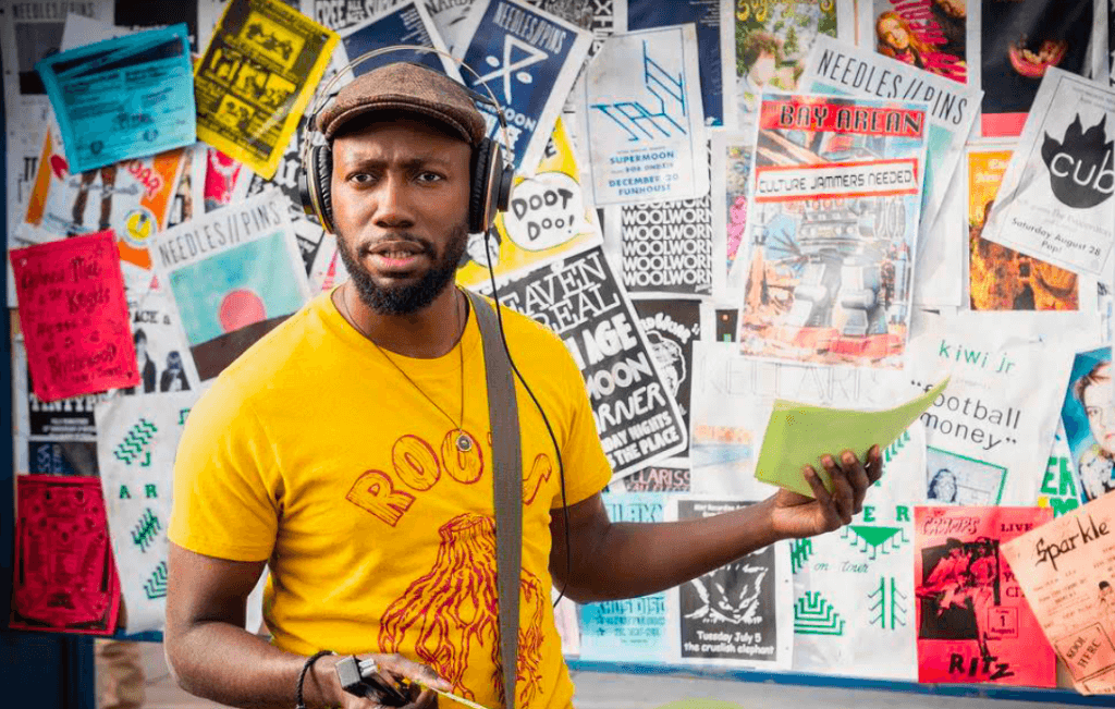 Still image from Woke actor Lamorne Morris stands in front of a bulletin board with a hat and headphones on
