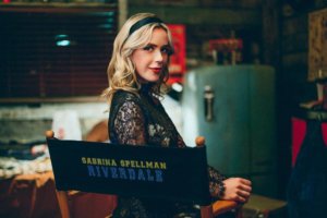 Kiernan shipka, a young thin blonde woman in her 20, sits sideways in a black director's chair. Her light blonde hair is held back by a black headband and she wears red lipstick and a black lace top. The chair reads "Riverdale" in blue collegiate-style letters and then Sabrina Spellman in yellow letters above it. 