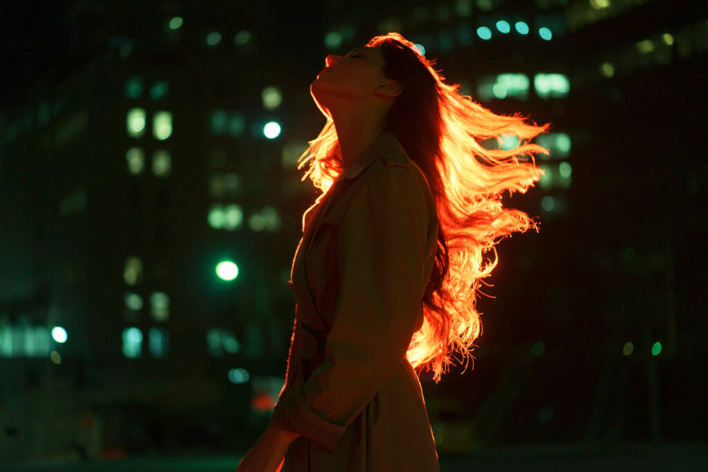 A female presenting person with long hair blowing in the wind against a cityscape at night