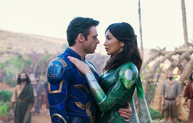 A white man with black hair in bright blue armor is standing close to an Asian woman with black hair in bright green armor. Her hands are on his chest and their gaze is romantic.