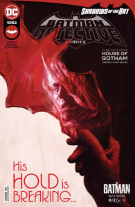 A hand holding a red batarang that is reflecting Batwoman's face