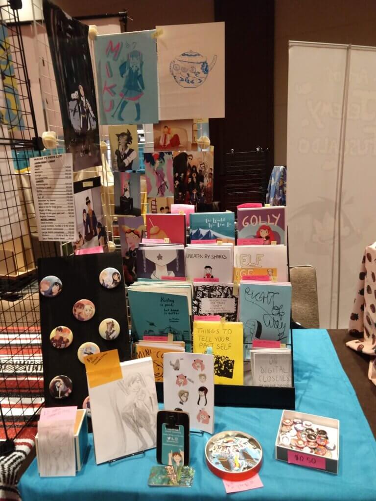 photo of my half-table setup with blue tablecloth, minicomics, prints, stickers and buttons displayed
