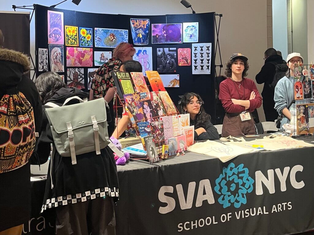 people stand and sit at a display table for the School of Visual Arts at MoCCA 2023 in this photo by Emily Lauer