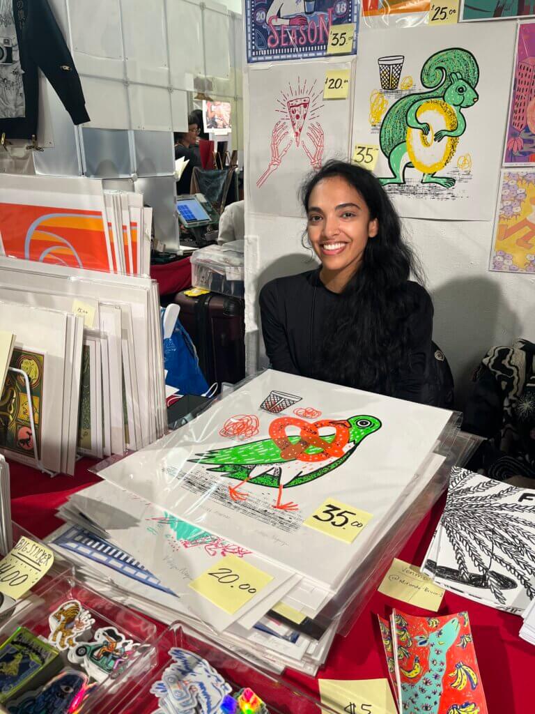 this photo by Emily Lauer shows artist Vidhya Nagarajan smiling sitting behind a table selling a sassy pigeon poster at MoCCA 2023.
