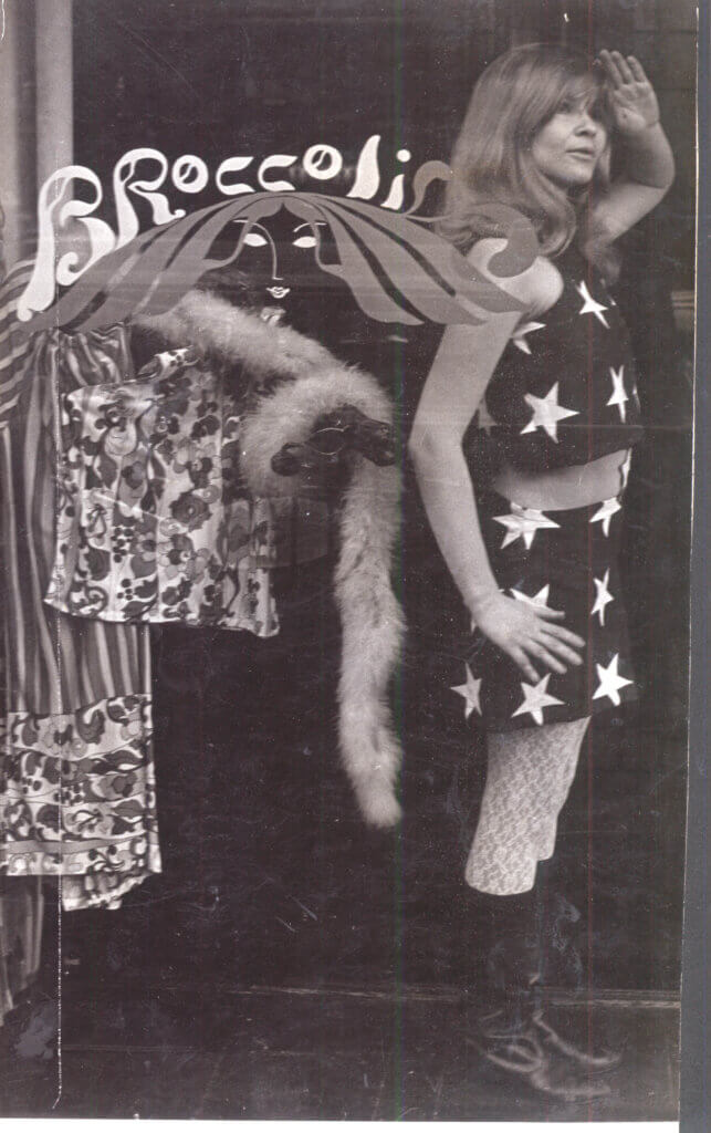 Trina Robbins is in her flag dress, saluting in the window of her boutique, Broccoli.