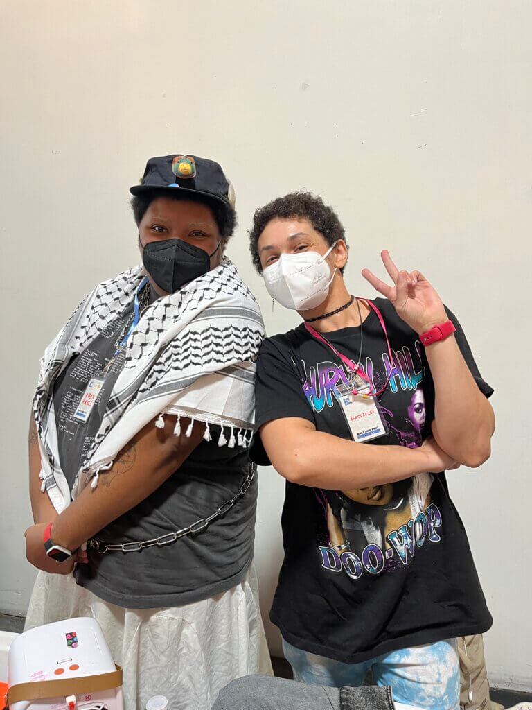 Two Black people one in a ball cap and one without stand behind a table at Black Zine Fest