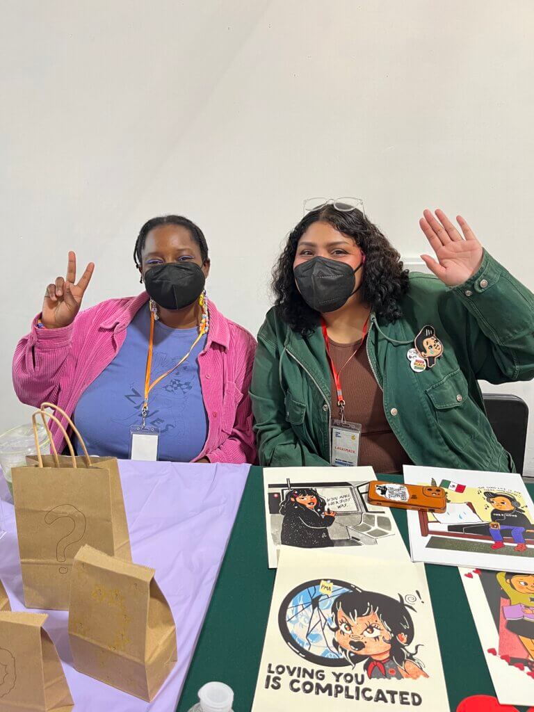 Two Black women sit behind a desk exhibiting zines at Black Zine Fest
