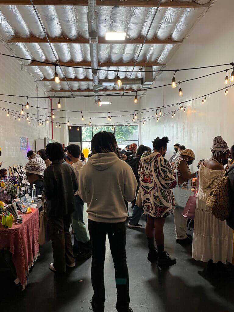 a crowd of people stands in a light airy room with high ceilings, where comics creators and zinesters exhibit at tables around them. This is Black Zine Fest the inaugural fest supporting Black zinesters.