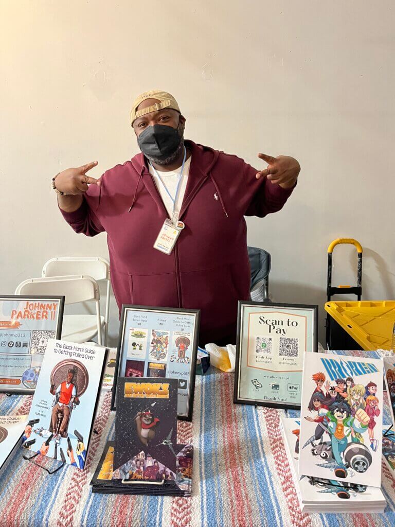 A Black man named Johnny Parker stands behind a table showcasing the books that he's worked on. 
