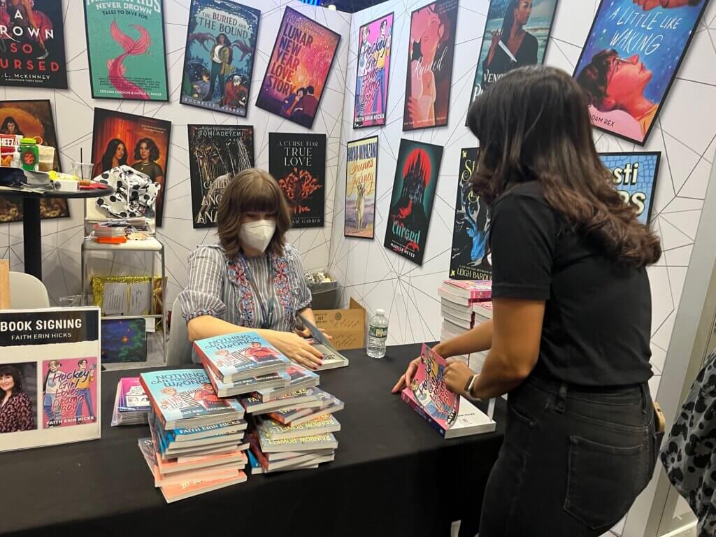Faith Erin Hicks, masked, signing for fans at the FIrst Second booth at NYCC 2023