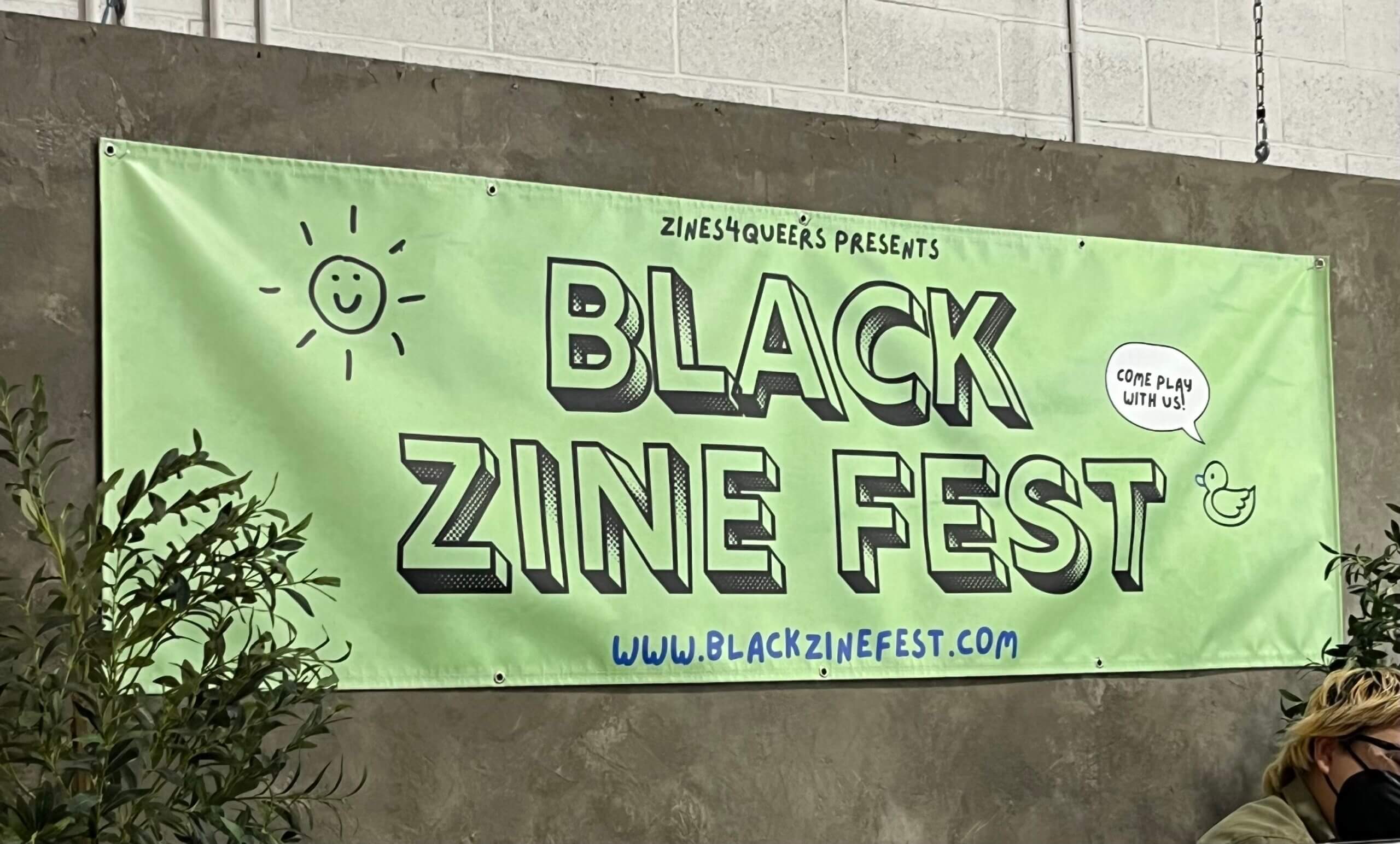 A green banner hangs on a grey wall with large text that reads Black Zine Fest in handwritten capitol letter font