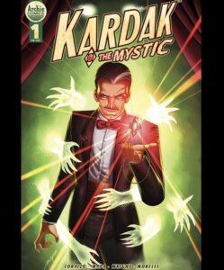 a man with dark hair in a dark tux stands upon a stage with a green backdrop; he's surrounded by many invisible hands which shoot out bolts of multicolored light.