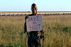 Still from the film Return of the Living Dead: Rave to the Grave. Shows a zombie holding a hitchhiking sign that reads "Rave or Bust".