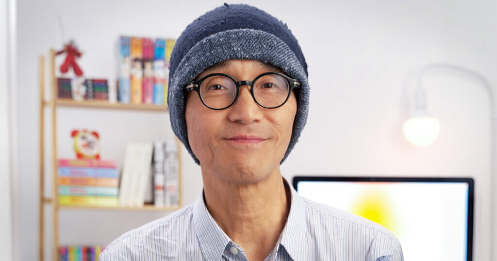 viz one shot program director Hisashi Sasaki, in knit cap and round glasses in front of bookshelves