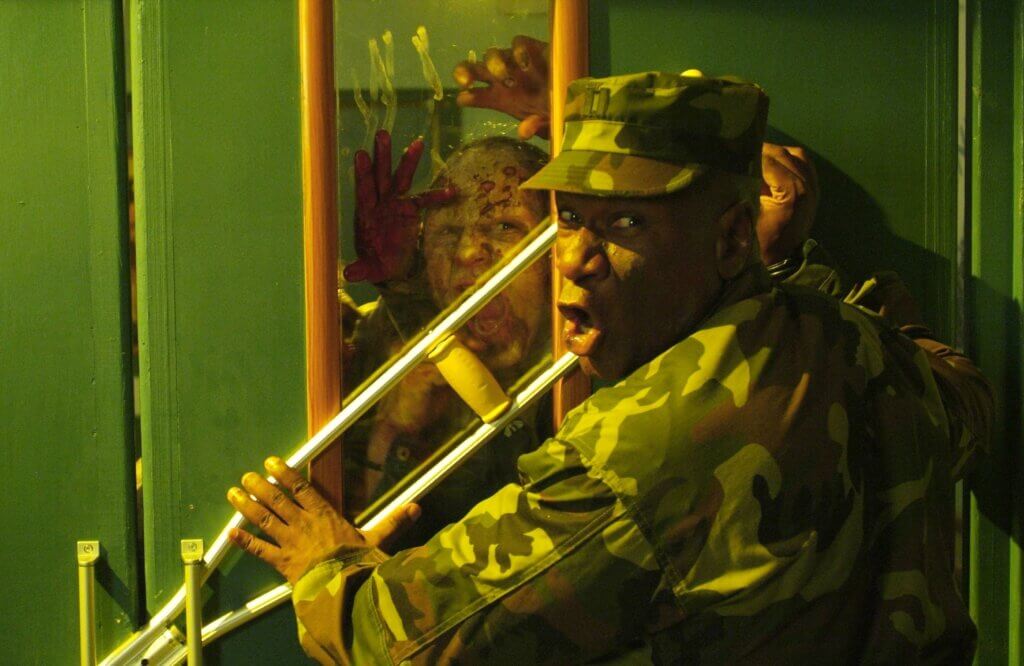 Still from the 2008 film Day of the Dead, showing a man in military uniform wedging a door shut with a crutch while a zombie claws at the window, all bathed in chartreuse hues.